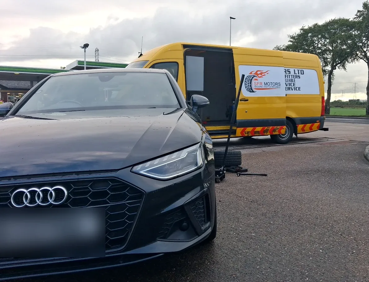 Black Audi at a service station with a trolley jack and a wheel on the ground beside a yellow SFR Motors mobile tyre van