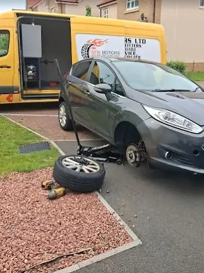Grey car with a front wheel removed and a trolley jack beside it, parked next to a yellow SFR Motors van with its side door open