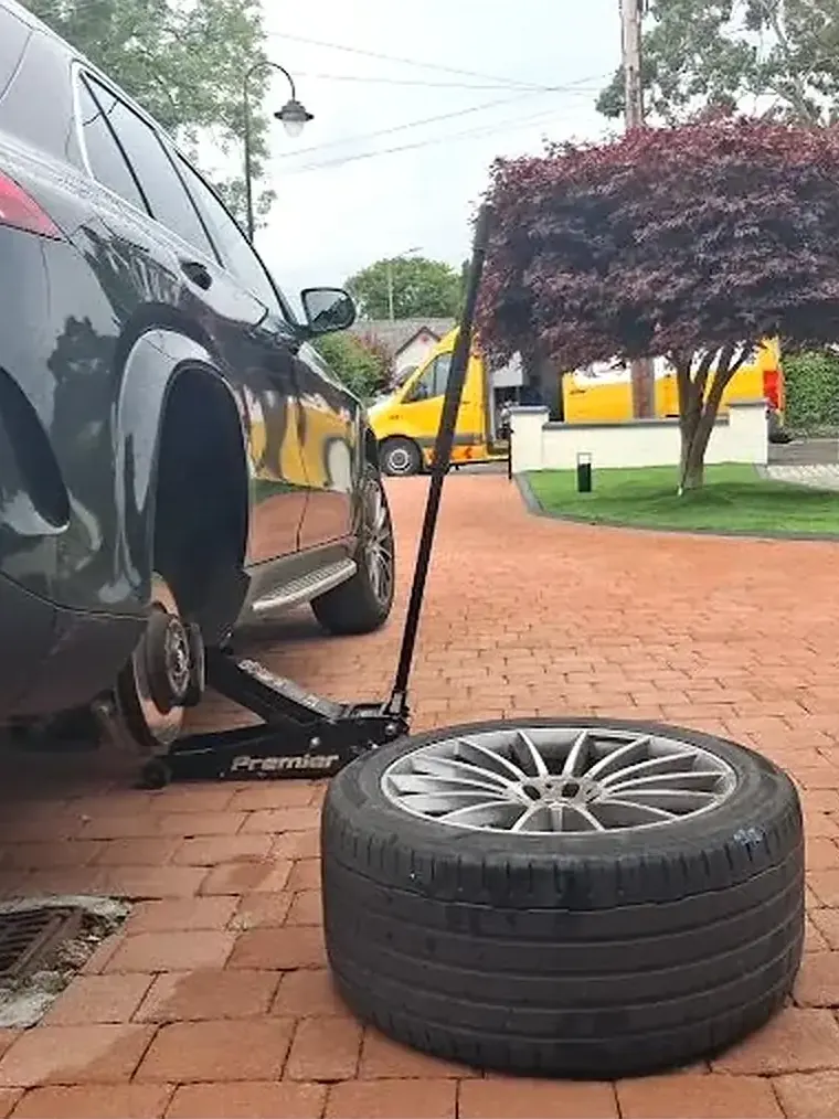 Black Mercedes SUV raised on a trolley jack with a wheel removed and standing on the block-paved driveway beside it