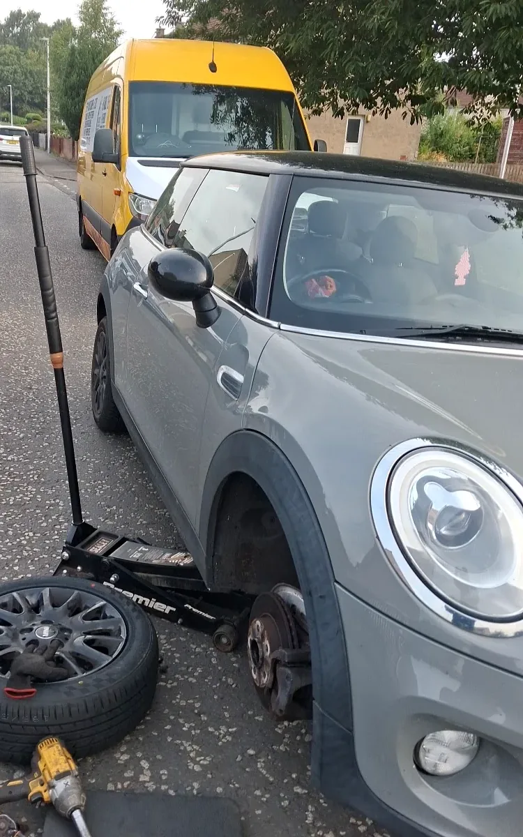 Grey Mini with its front wheel removed and a trolley jack, wheel and impact wrench beside it on a residential street, with a yellow van parked behind